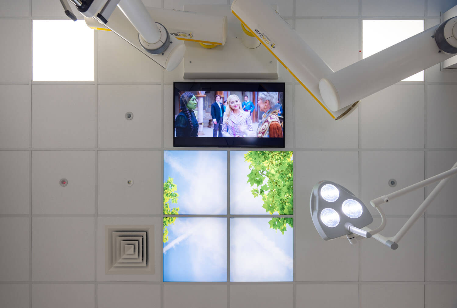 Ceiling view a patient sees from the treatment bed, with backlit sky panel, surgical light and TV
